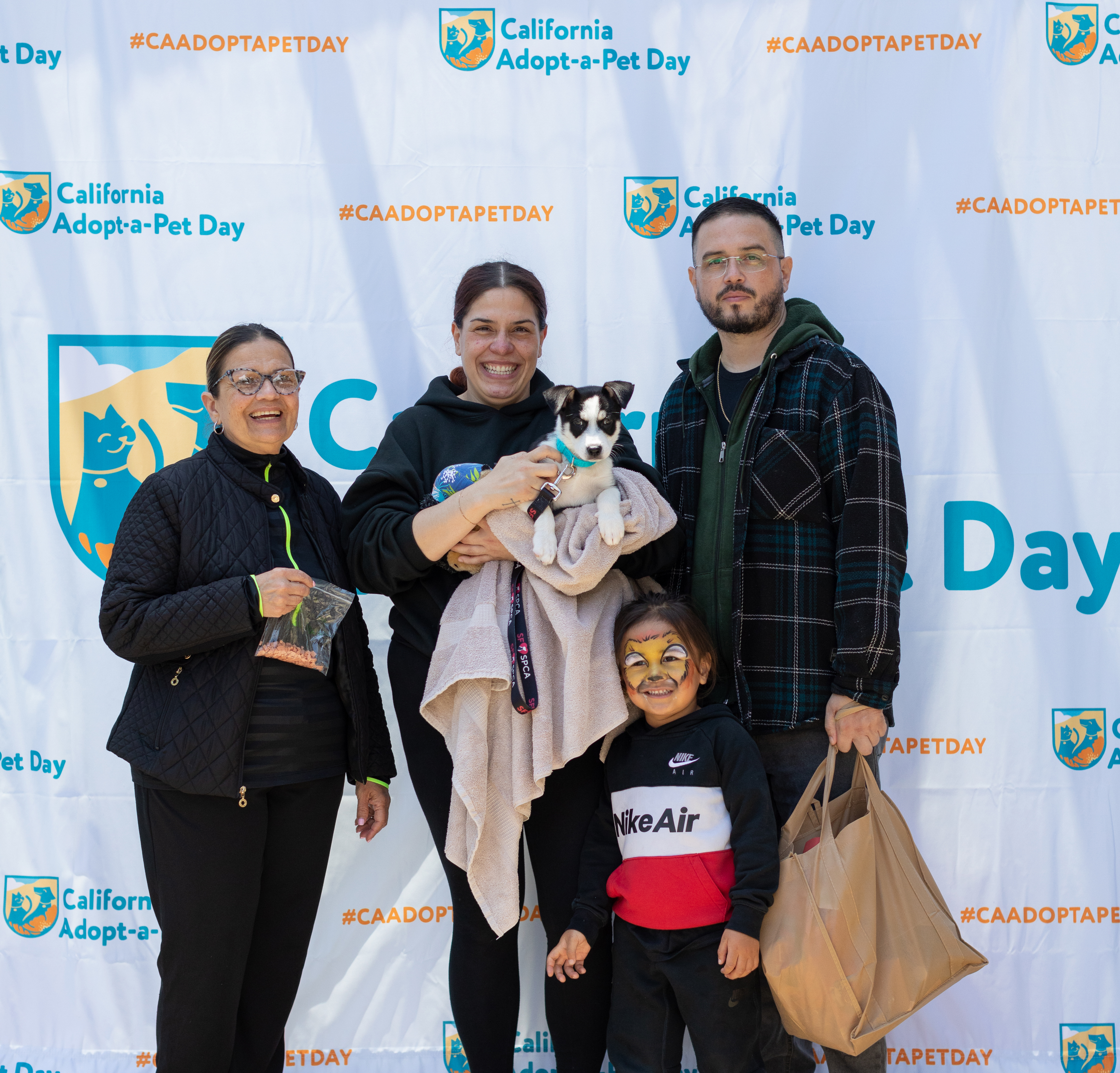 “Two women and one man posing with a small puppy and a young child in front of a California Adopt-a-Pet Day backdrop, smiling after an adoption event.”