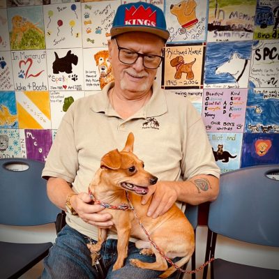 Older man wearing glasses and a baseball cap sitting indoors while holding a small tan dog on his lap, with a wall of memorial pet tiles behind them