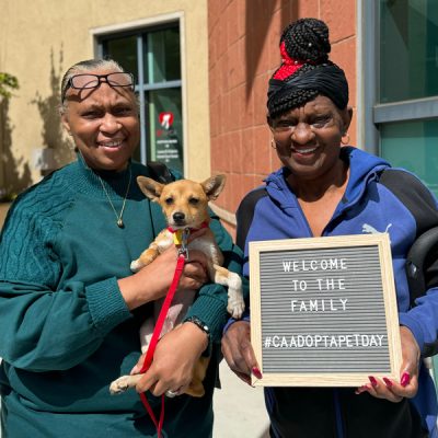 Two women smiling outdoors, one holding a small tan dog on a red leash and the other holding a sign that reads ‘Welcome to the family #CAAdoptAPetDay.’