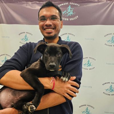 Man smiling while holding a black puppy in front of a Humane Society Silicon Valley backdrop